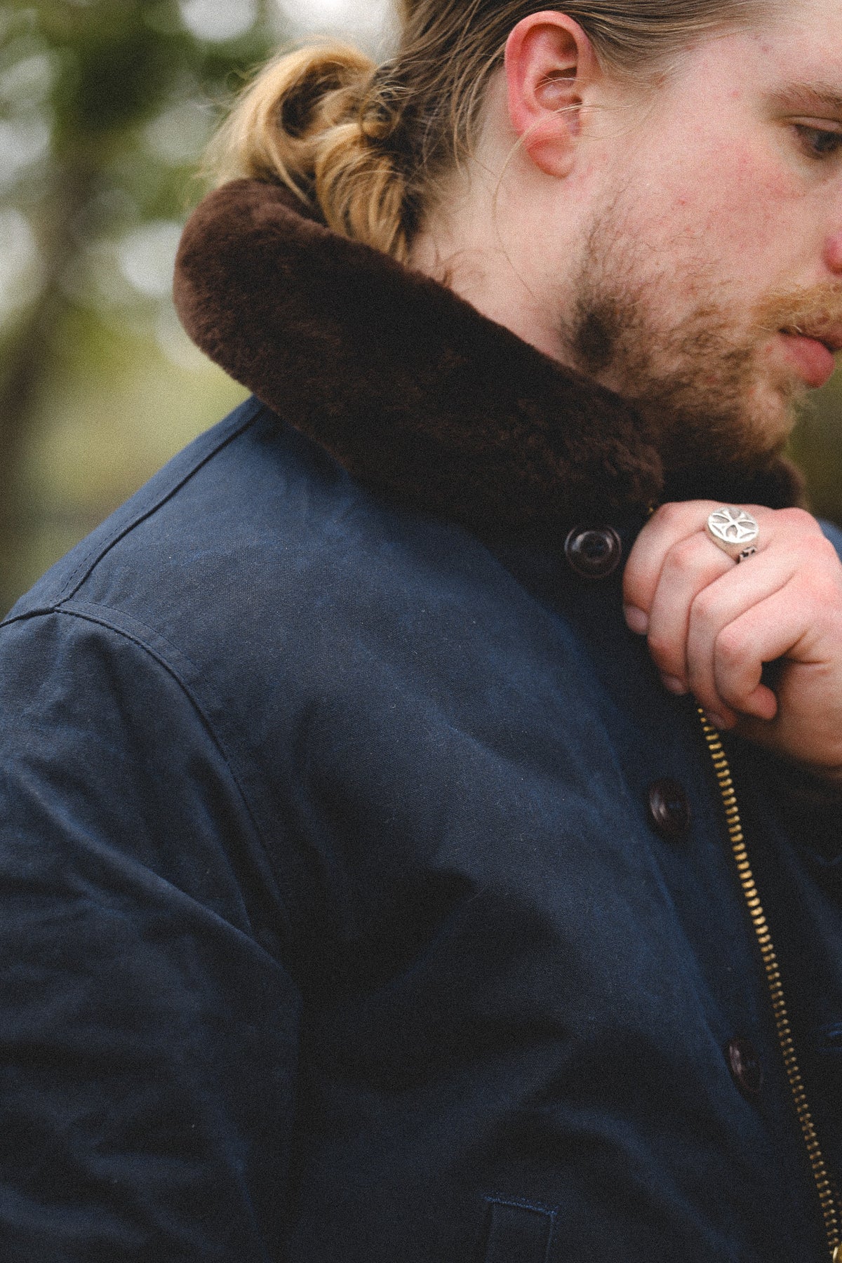 A close-up of a man wearing a navy blue waxed canvas jacket with a dark brown faux-fur collar, outdoors.