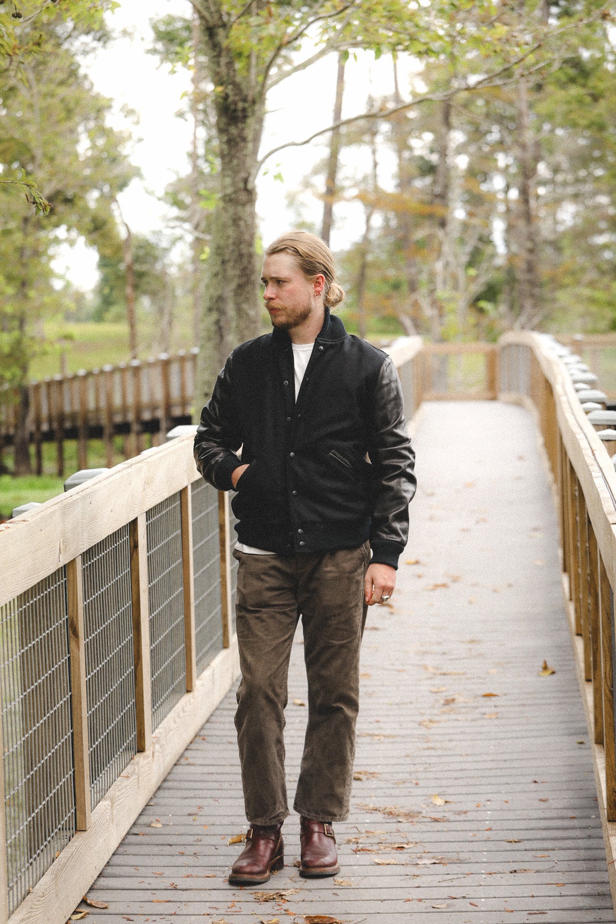 A man wearing a black leather bomber jacket, brown corduroy pants, and brown boots walks across a wooden boardwalk surrounded by trees.