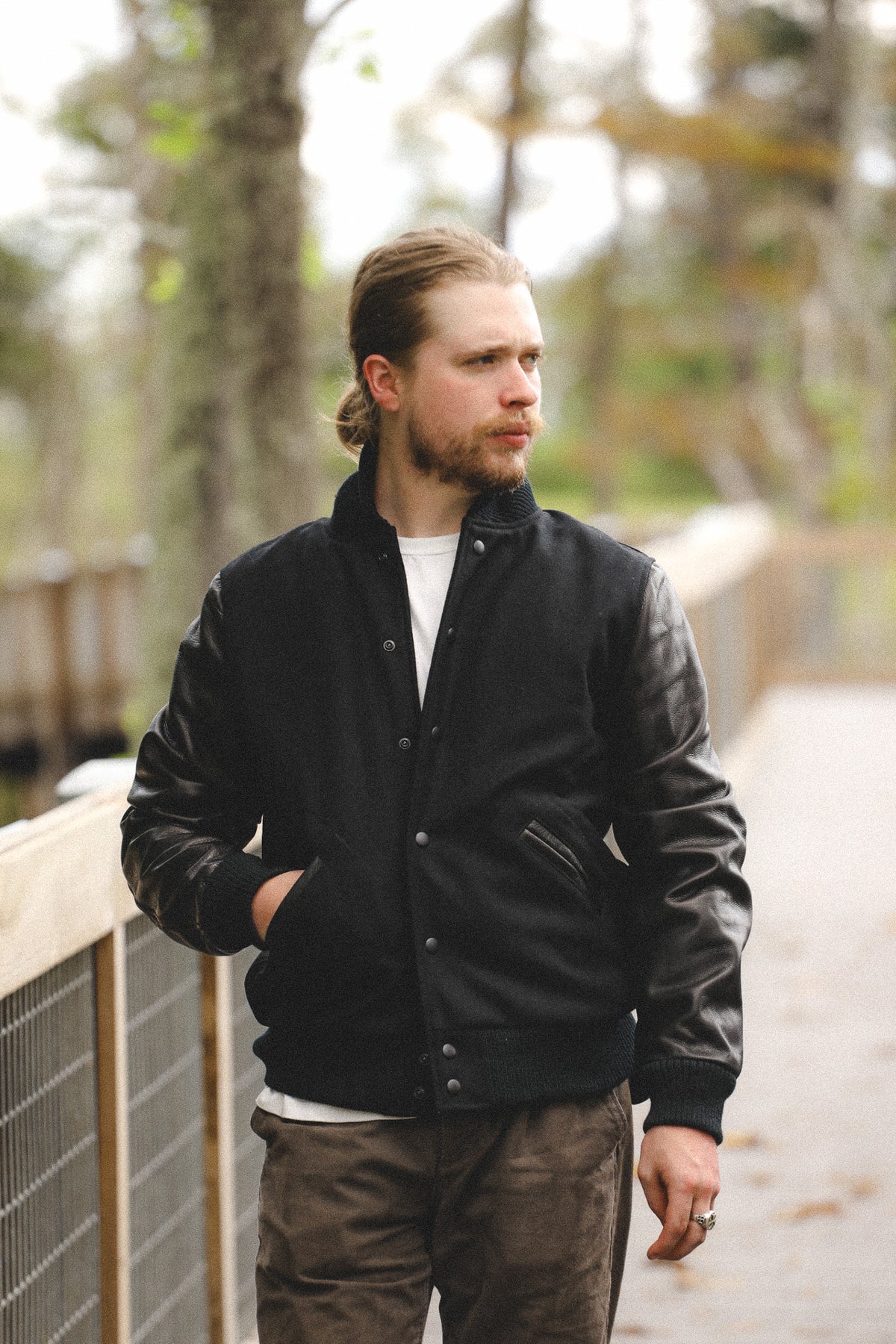 A man wearing a black varsity-style jacket with dark leather sleeves, standing on an outdoor wooden boardwalk path.