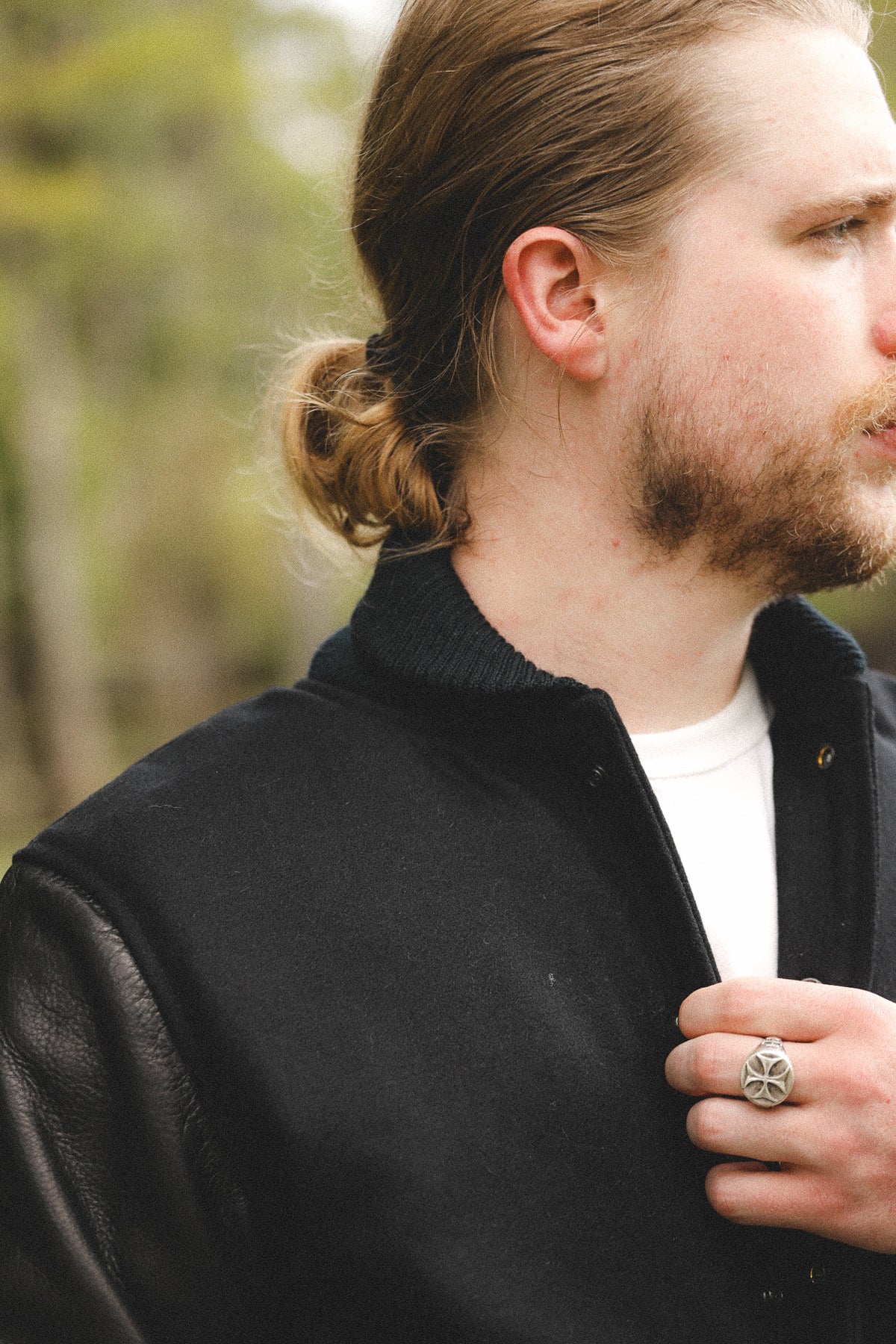 A close-up side profile of a man with a beard and long hair in a low bun, wearing a black leather-look jacket over a white t-shirt, with a silver ring on his finger, set against a blurred outdoor background.
