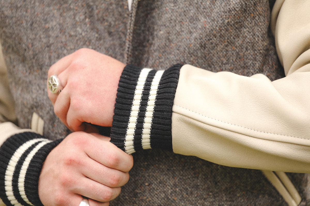 Close-up of a person's hands adjusting the cuff of a cream leather and black-and-white striped knit varsity jacket.