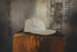 A light beige textured baseball cap with a flat visor, resting on a rustic wooden block against a blurred gray concrete background.
