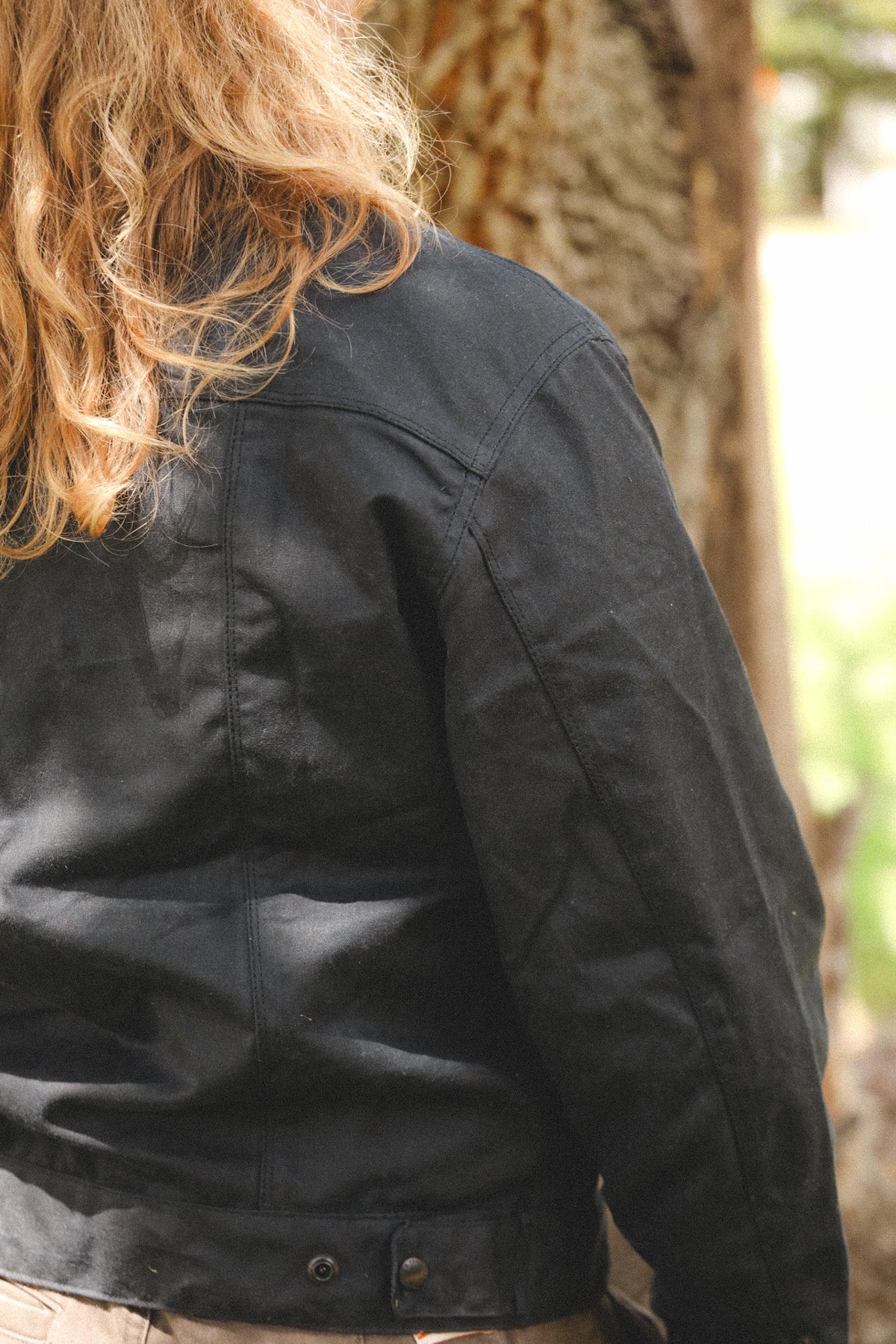 A close-up back view of a person wearing a black jacket outdoors in front of a tree trunk.