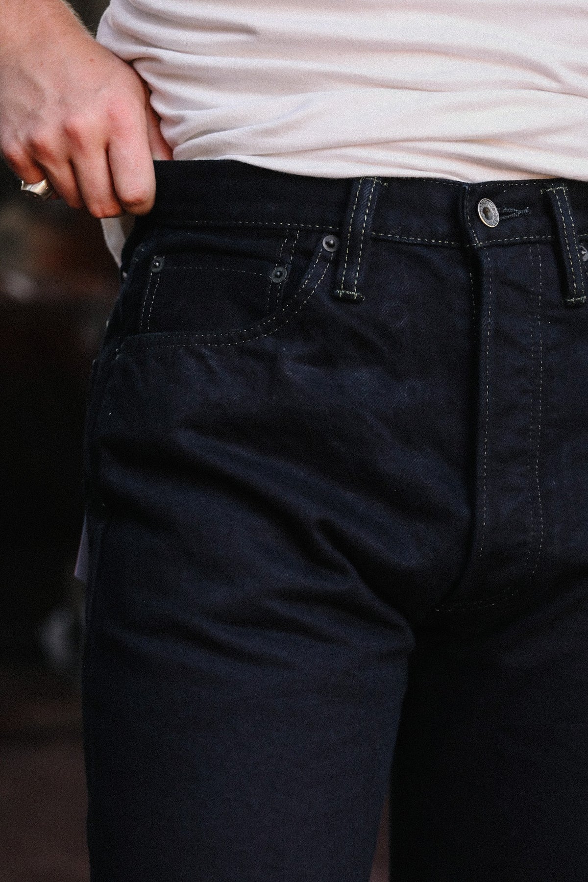Close-up of a person wearing dark indigo denim jeans with a light-colored t-shirt, showing the waistband, button, and front pockets.