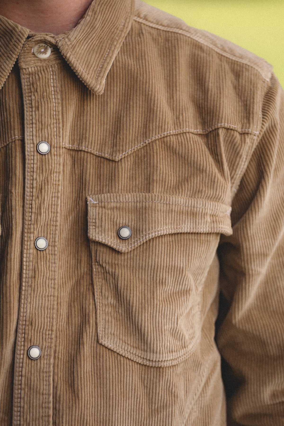 Close-up of a tan corduroy button-up shirt worn by a person, showing the chest pocket, buttons, and collar against a blurred yellow-green background.
