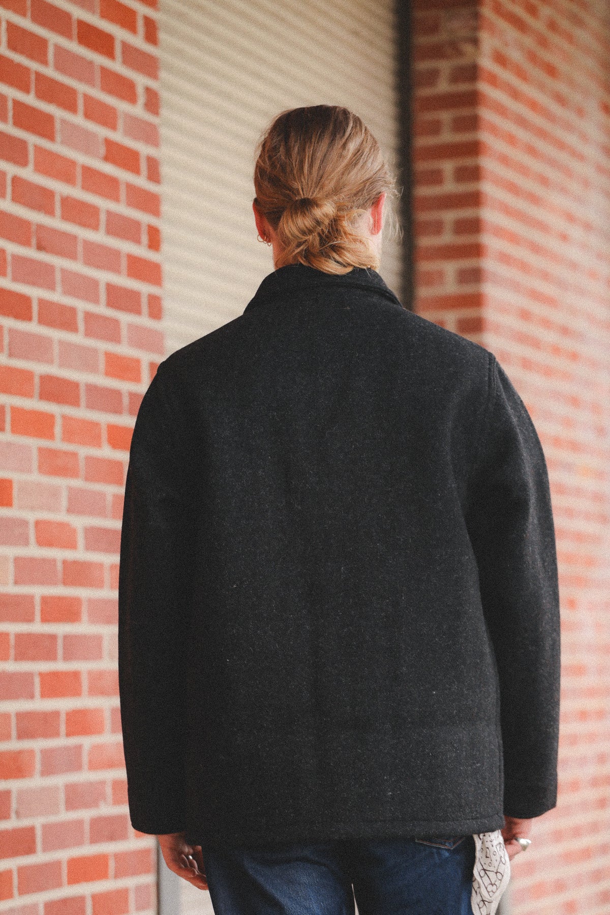 A person viewed from behind wearing a dark charcoal gray wool-blend coat, standing in front of a red brick wall and a metal roll-up door.