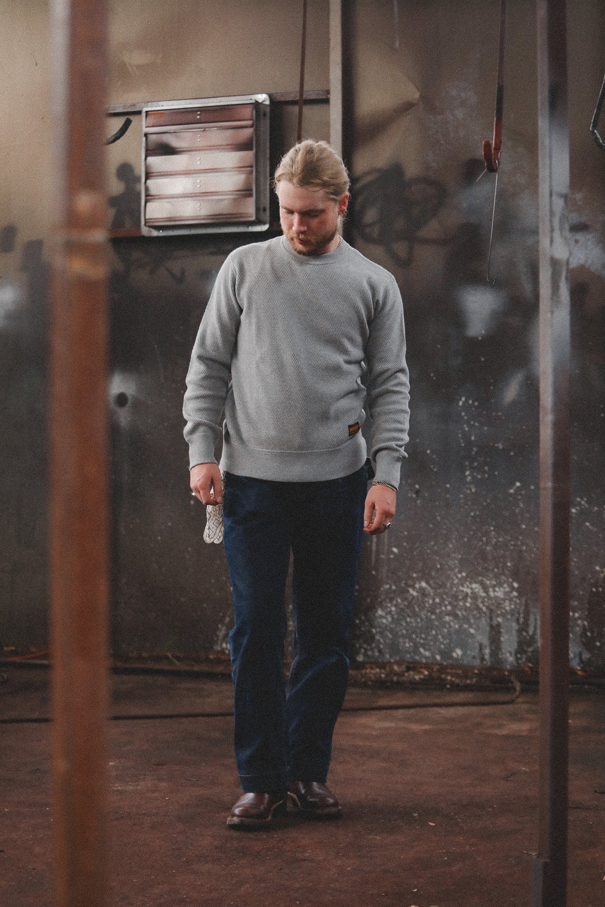 A man wearing a light gray crewneck sweatshirt, dark blue jeans, and brown shoes standing in a gritty, industrial room with graffiti on the wall.