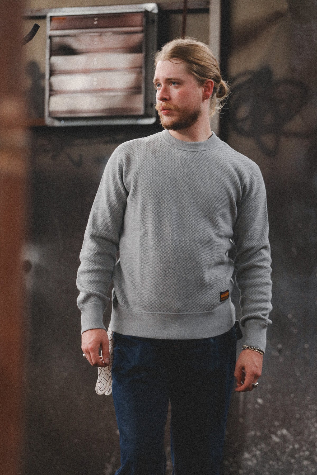 A man with blonde hair tied back wears a light gray crewneck sweatshirt and dark blue jeans, standing against a dark, textured urban wall.