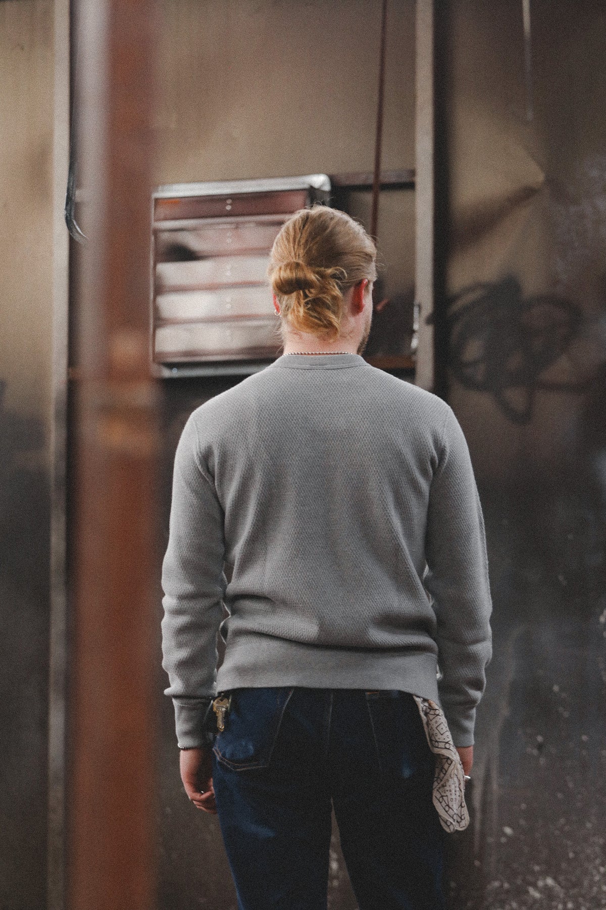 A person viewed from behind wearing a gray long-sleeve crewneck sweater and dark blue jeans, standing in a dimly lit urban alleyway.