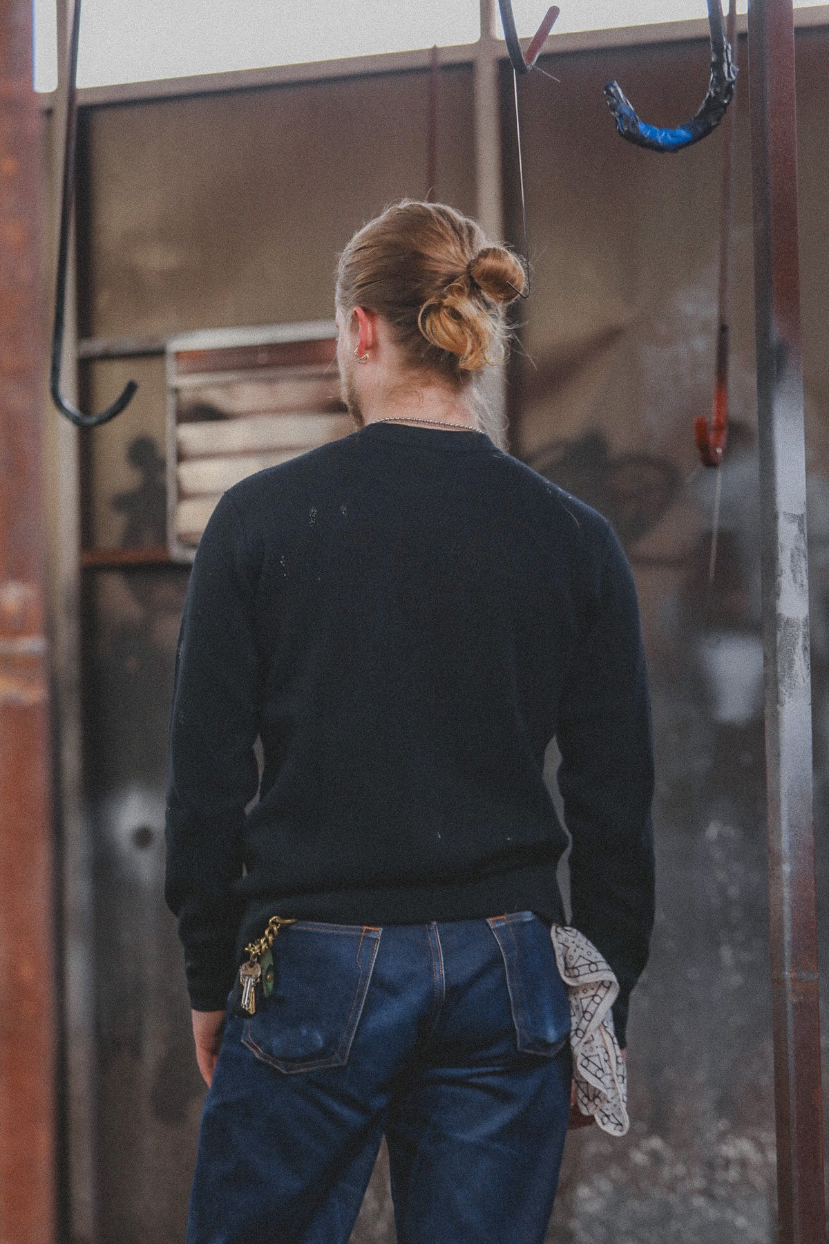 A person viewed from behind wearing a black long-sleeve crewneck sweater and blue jeans, standing in front of a rustic metal wall with hanging hooks.