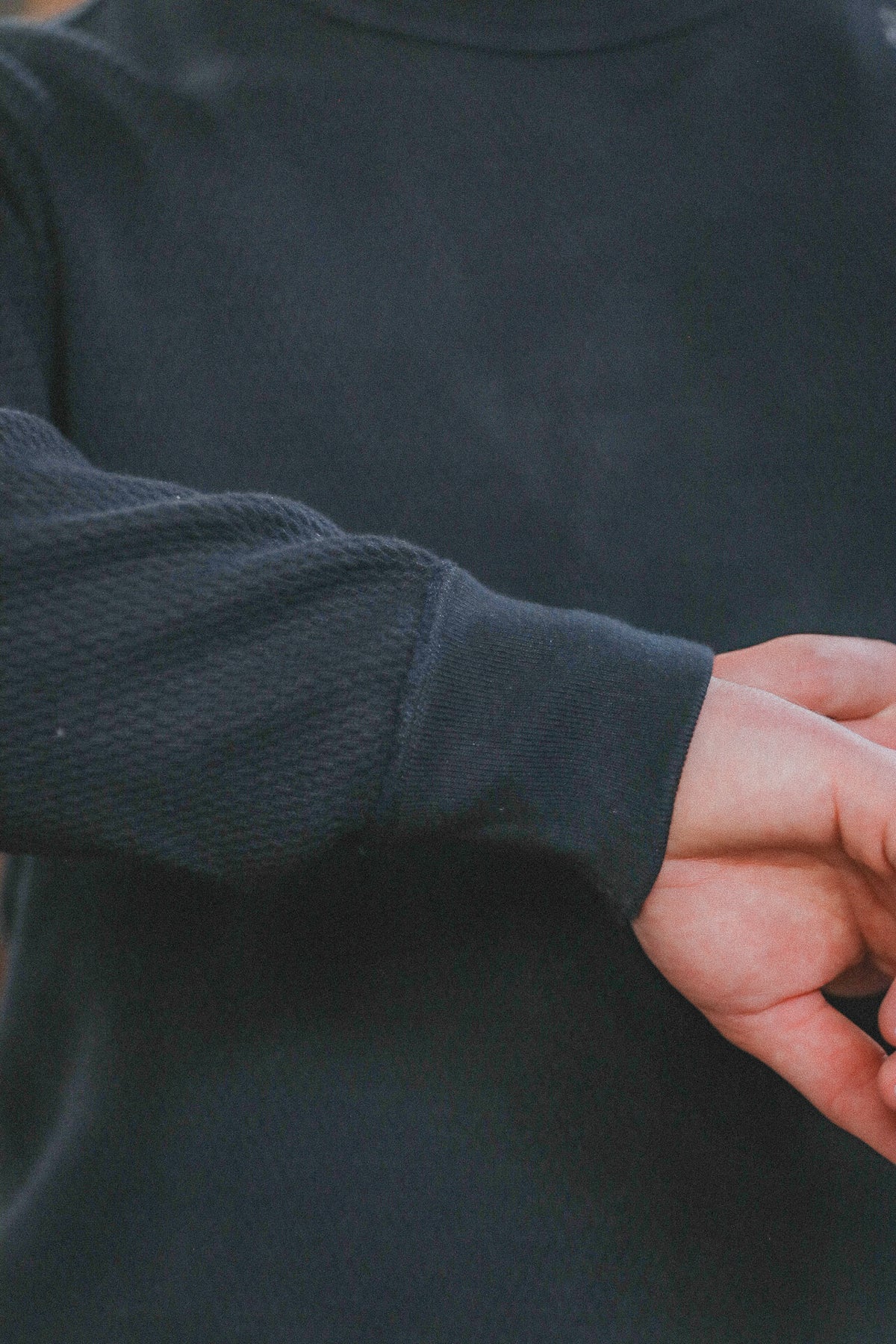 Close-up of a person wearing a dark charcoal gray long-sleeve knit top, showing the textured fabric and ribbed cuff with a hand adjusting or touching the sleeve.