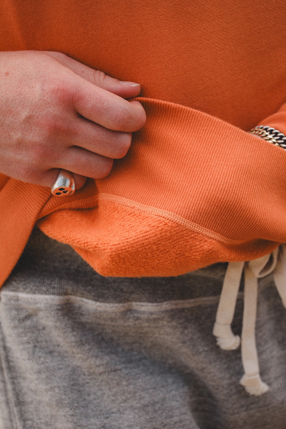 Close-up of a person wearing an orange ribbed knit top and gray drawstring sweatpants, with a hand pulling the hem of the orange garment.