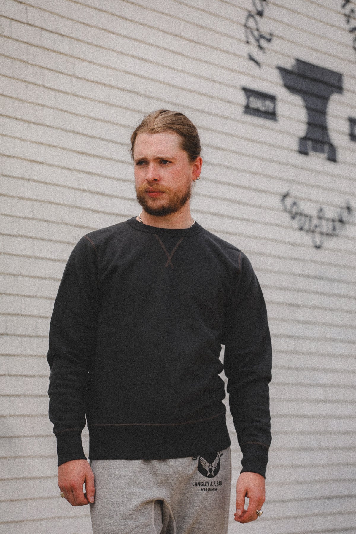 A man wearing a dark charcoal crewneck sweatshirt and light gray sweatpants, standing against a white brick wall with black graffiti.