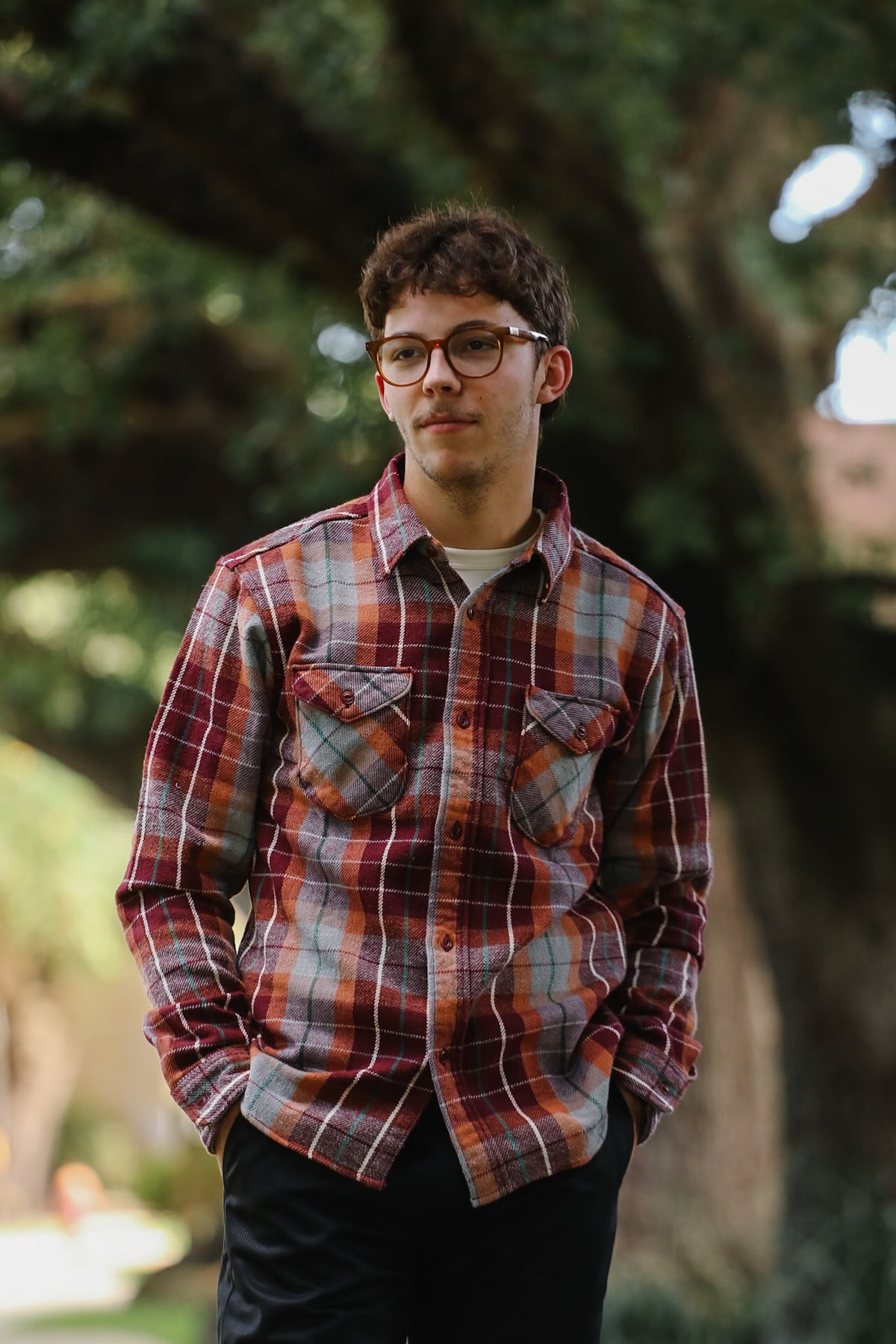 A young man wearing a red, orange, and gray plaid button-down flannel shirt with glasses, standing outdoors in front of a large, blurred tree.