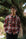 A young man wearing a red, orange, and gray plaid button-down flannel shirt with glasses, standing outdoors in front of a large, blurred tree.