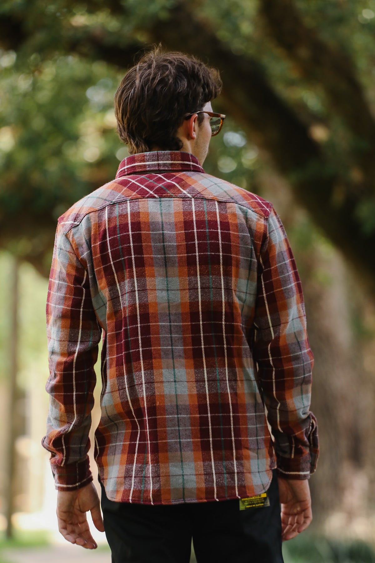 A man viewed from behind wearing a long-sleeve plaid button-up shirt in rust orange, red, and gray with thin white lines, paired with black pants, set against a blurred outdoor background of trees.