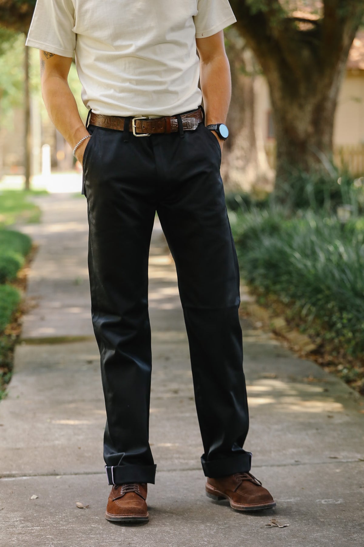 A person standing on a sidewalk outdoors wearing dark navy trousers, a cream short-sleeve shirt, a brown belt, and brown shoes.