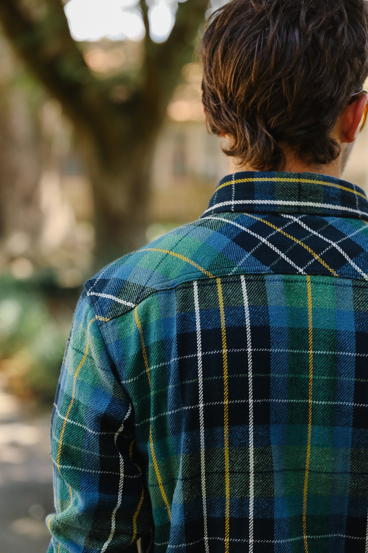 A man viewed from behind wearing a green and blue plaid flannel shirt with yellow and white stripes, set against a blurred outdoor background.