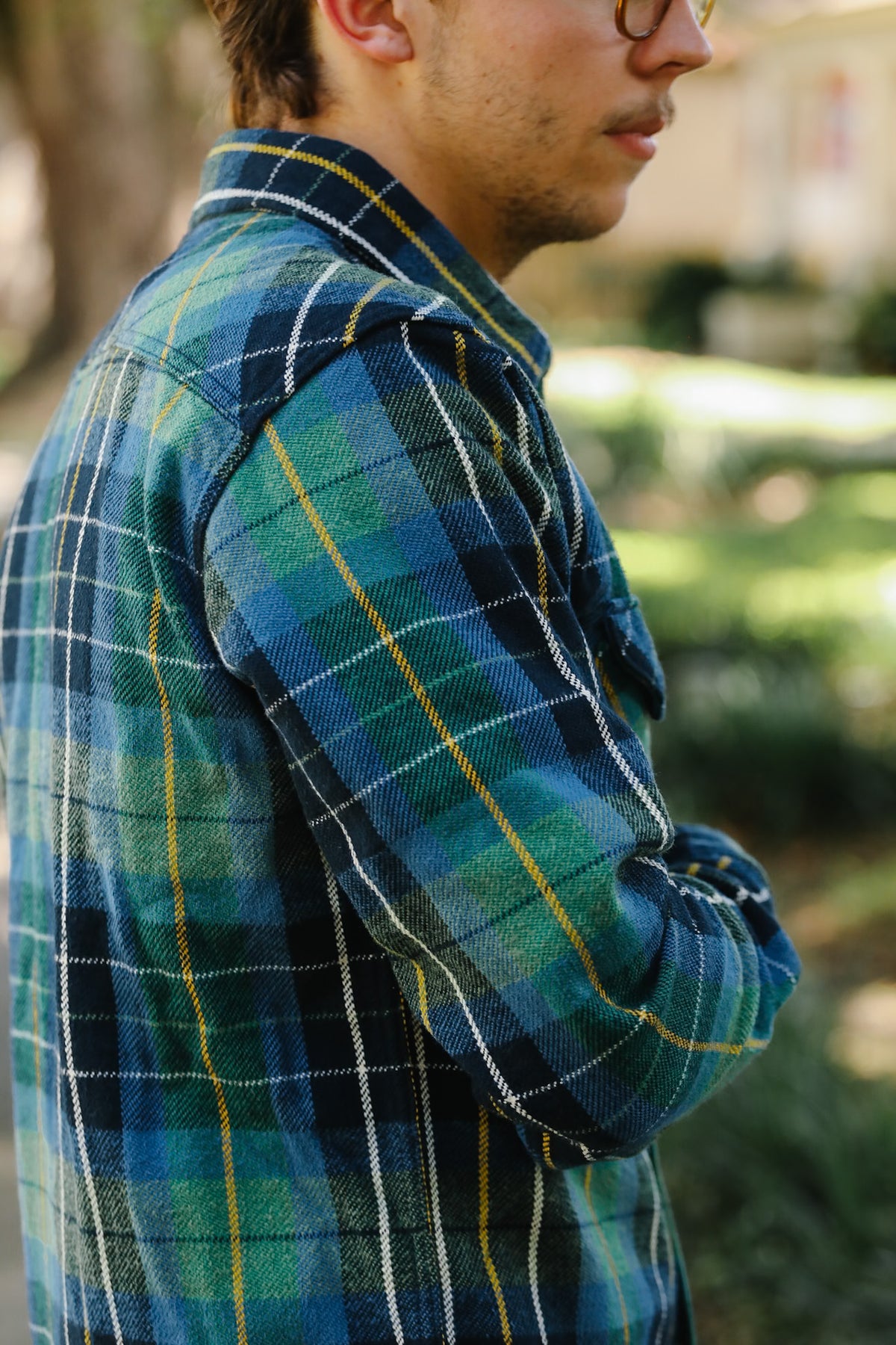 A man wearing a blue and green plaid flannel shirt with yellow and white stripes, photographed outdoors with a blurred park background.