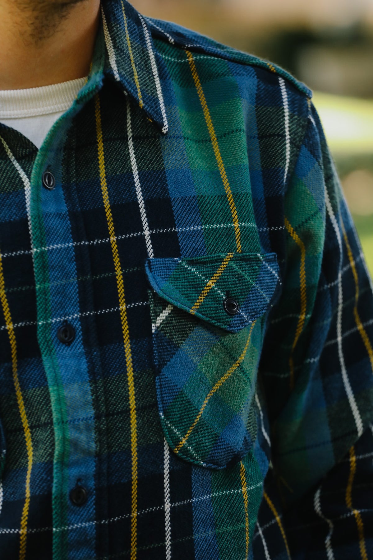 Close-up of a men's long-sleeve plaid flannel shirt in blue, green, and yellow, worn over a white undershirt.