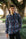 A young man wearing round glasses and a blue, red, and white plaid button-down shirt, standing outdoors on a paved path with trees in the background.