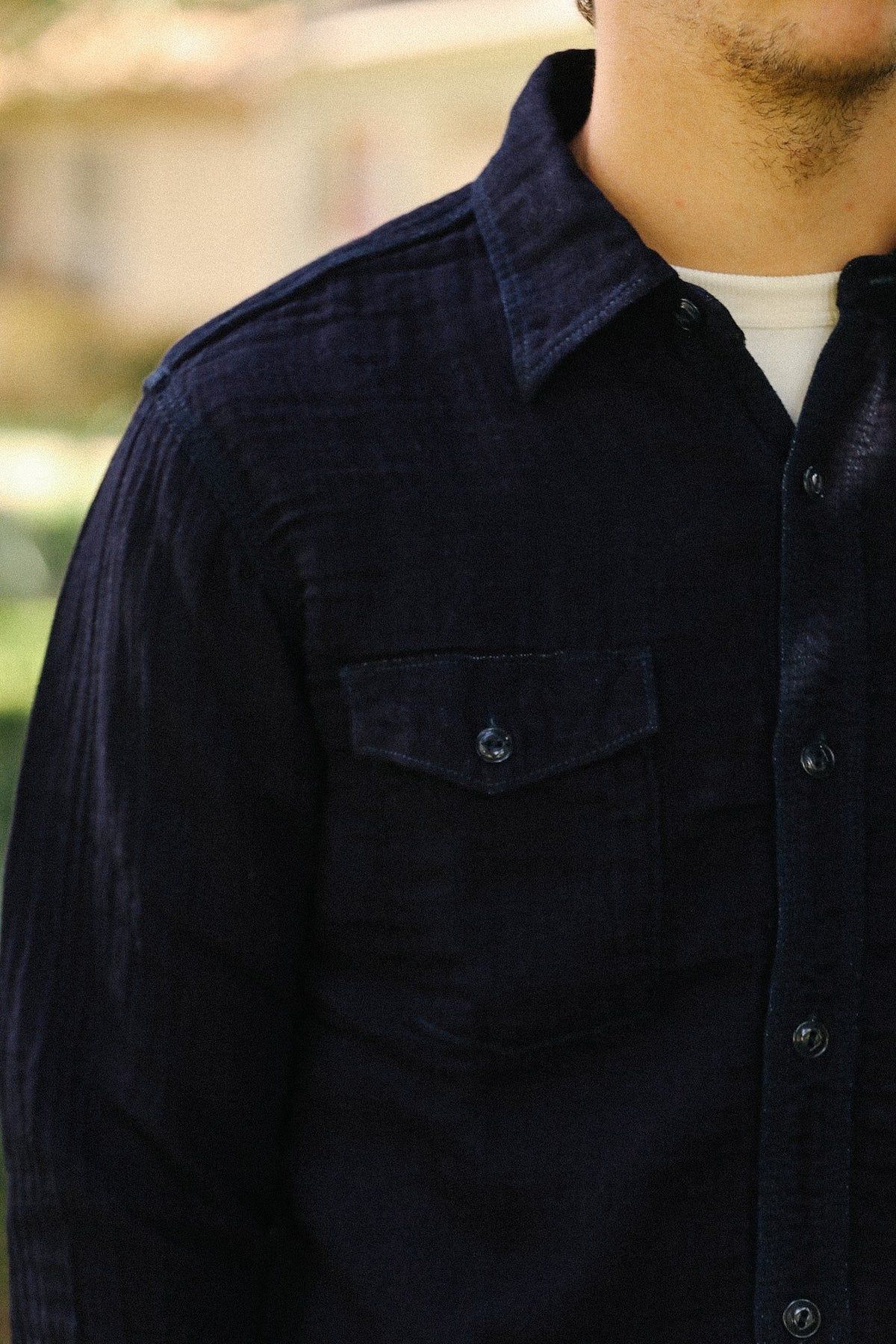 Close-up of a man wearing a dark navy textured button-up shirt over a white undershirt, with a blurred outdoor background.