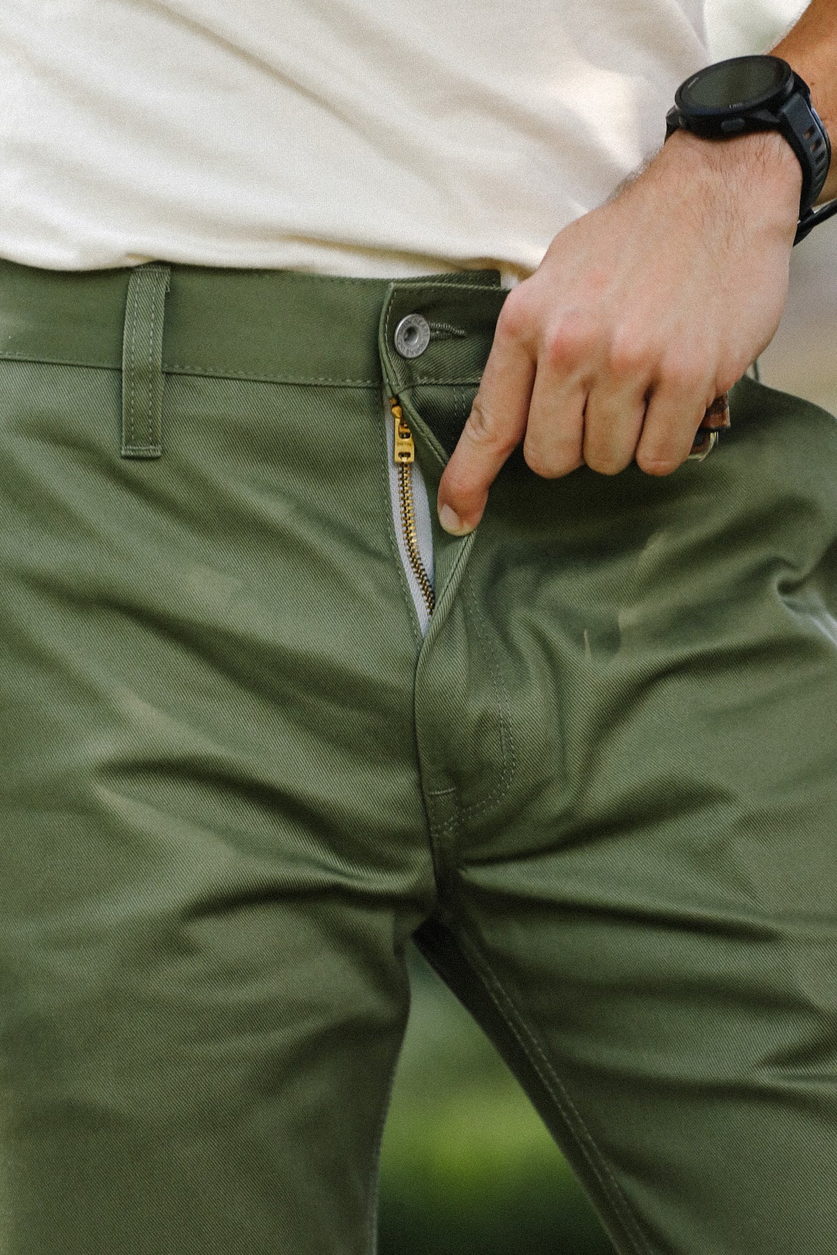 Close-up of a person wearing olive green trousers, pulling the zipper open with their hand. The person is also wearing a white shirt and a black smartwatch.