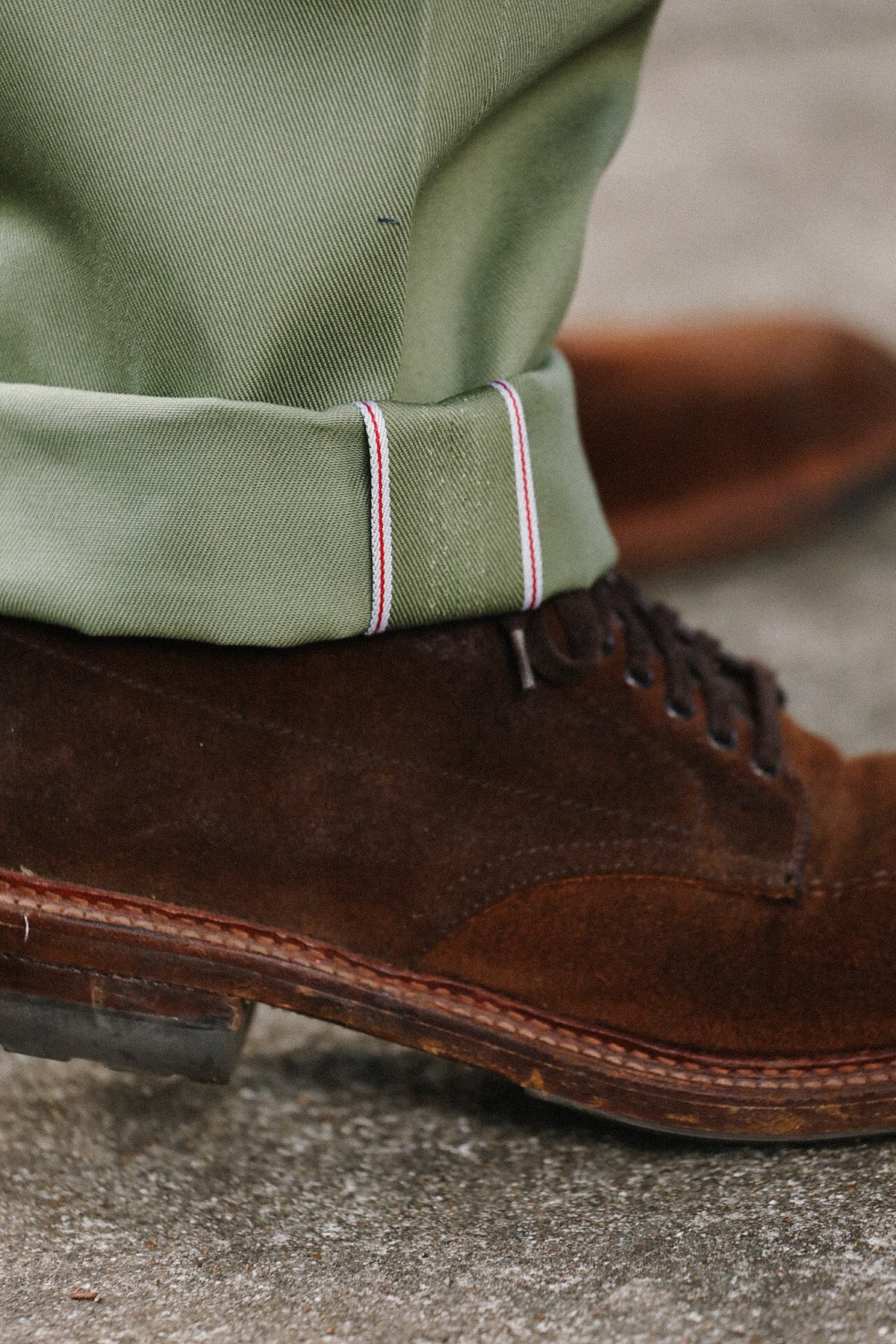 A close-up shot of a person's foot wearing a brown suede lace-up shoe and an olive green trouser leg with a striped cuff, standing on a gray concrete surface.