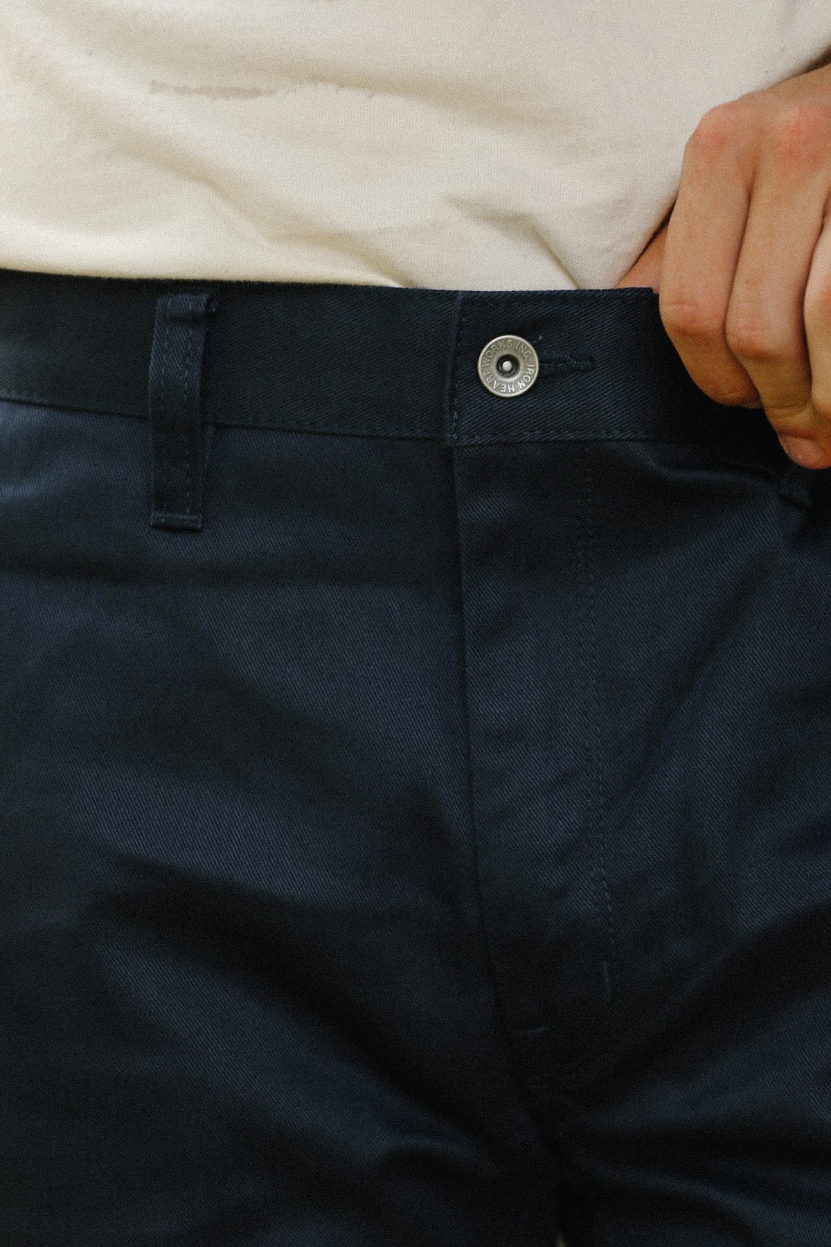 Close-up of a person wearing dark navy trousers, showing the waistband with a silver button, belt loops, and part of a light-colored shirt, with a hand pulling the fabric slightly.