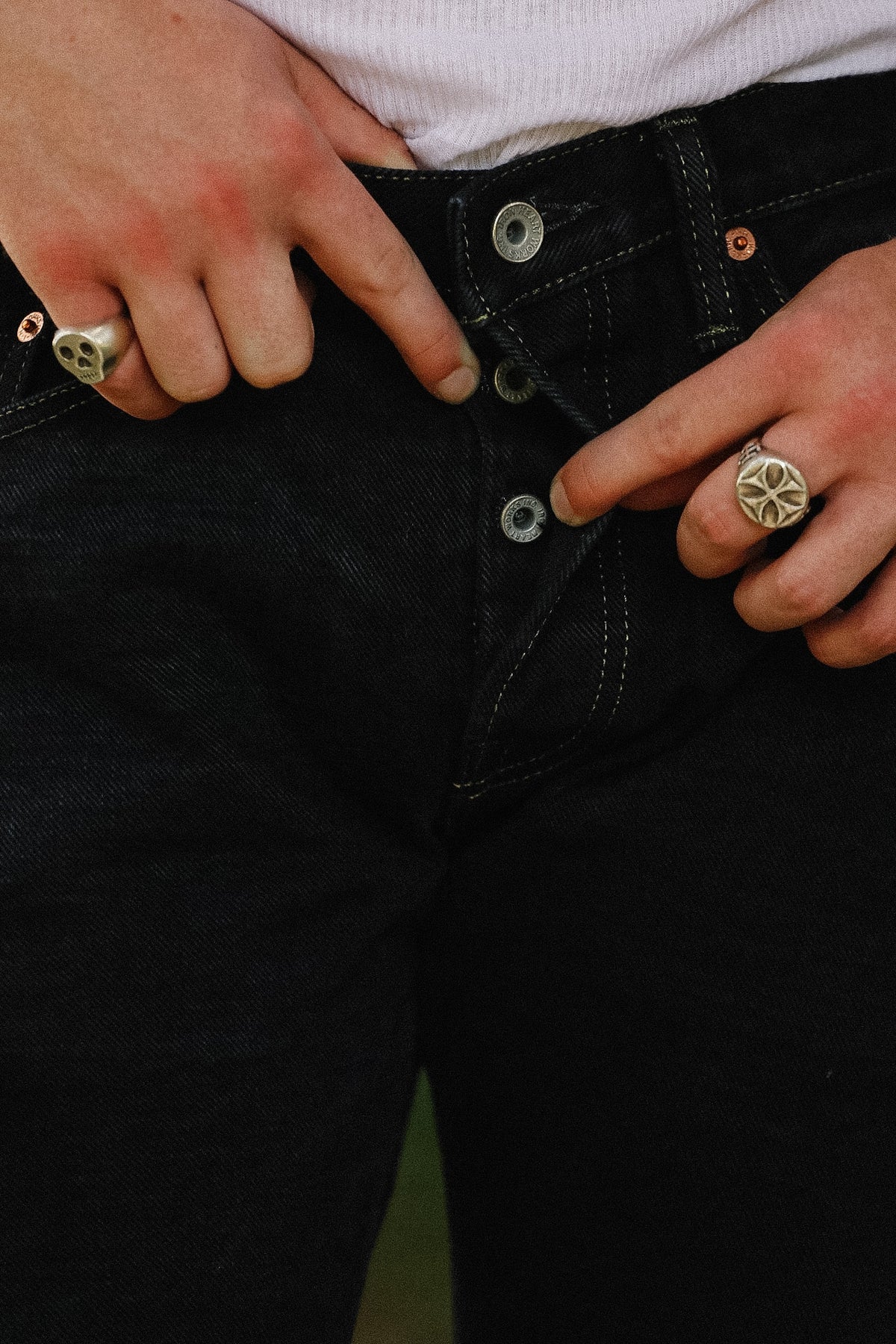 Close-up of a person wearing black denim jeans, showing the front button closure with silver metal buttons, contrast stitching, and hands adjusting the waistband. The person is also wearing silver rings.