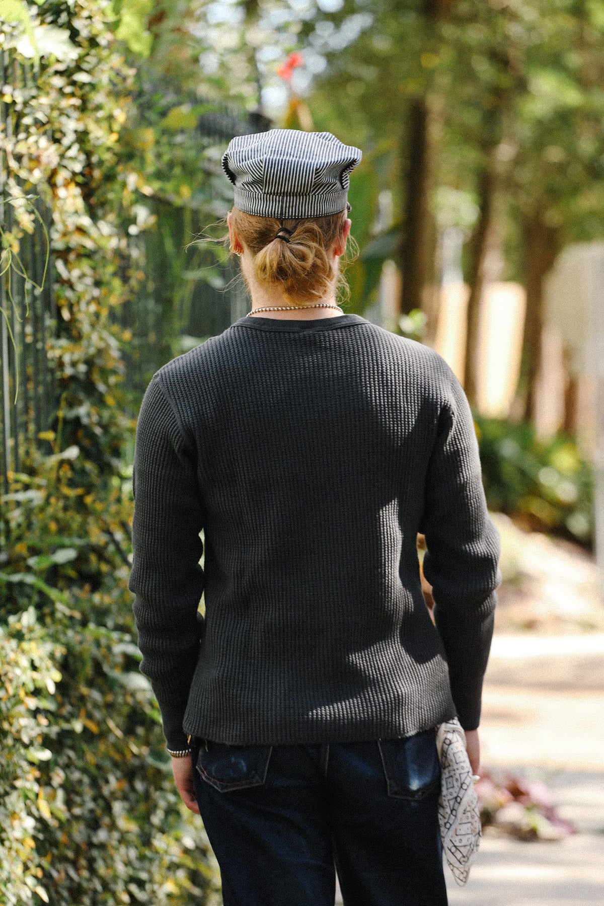 A person viewed from behind wearing a dark charcoal gray ribbed knit sweater, a gray and white striped flat cap, and dark blue jeans, walking outdoors on a sunlit path lined with green foliage.