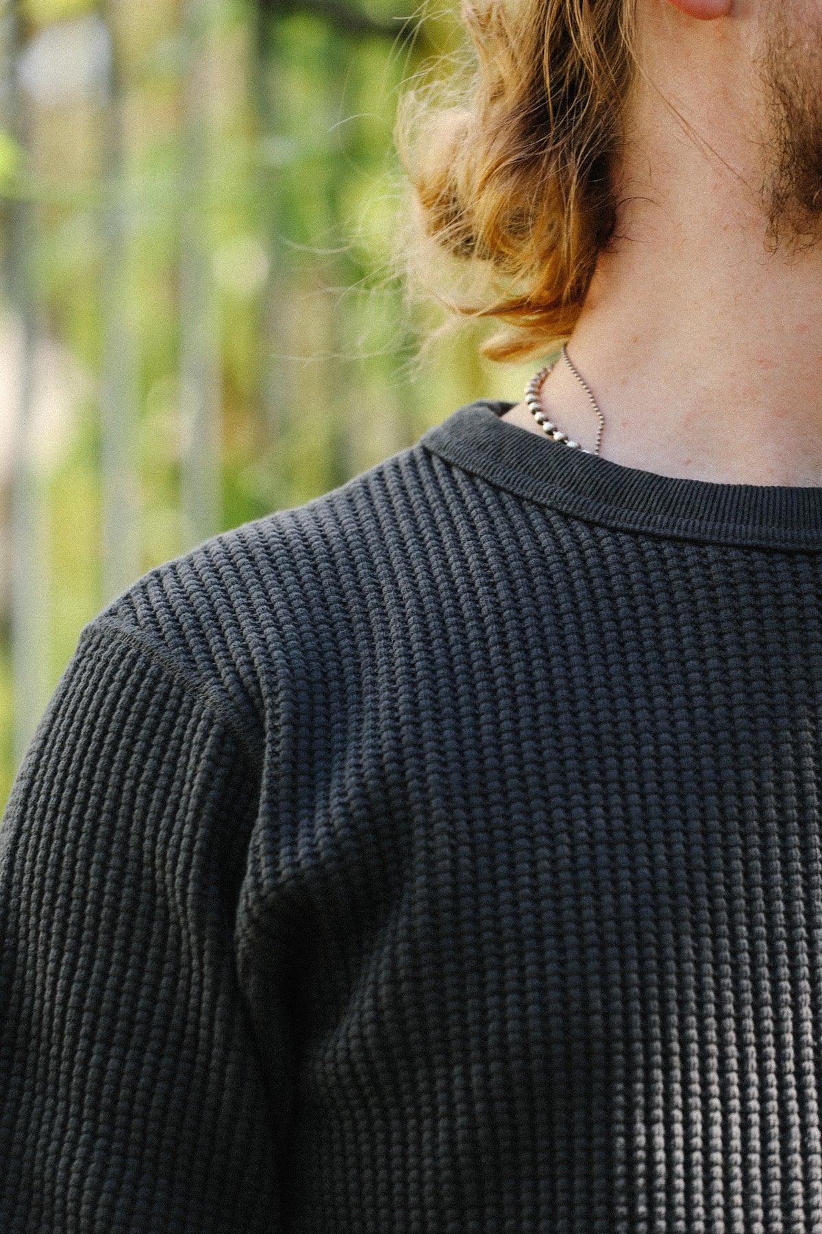 Close-up of a person wearing a dark charcoal gray waffle-knit crewneck sweater, with long light brown hair and a blurred outdoor background.