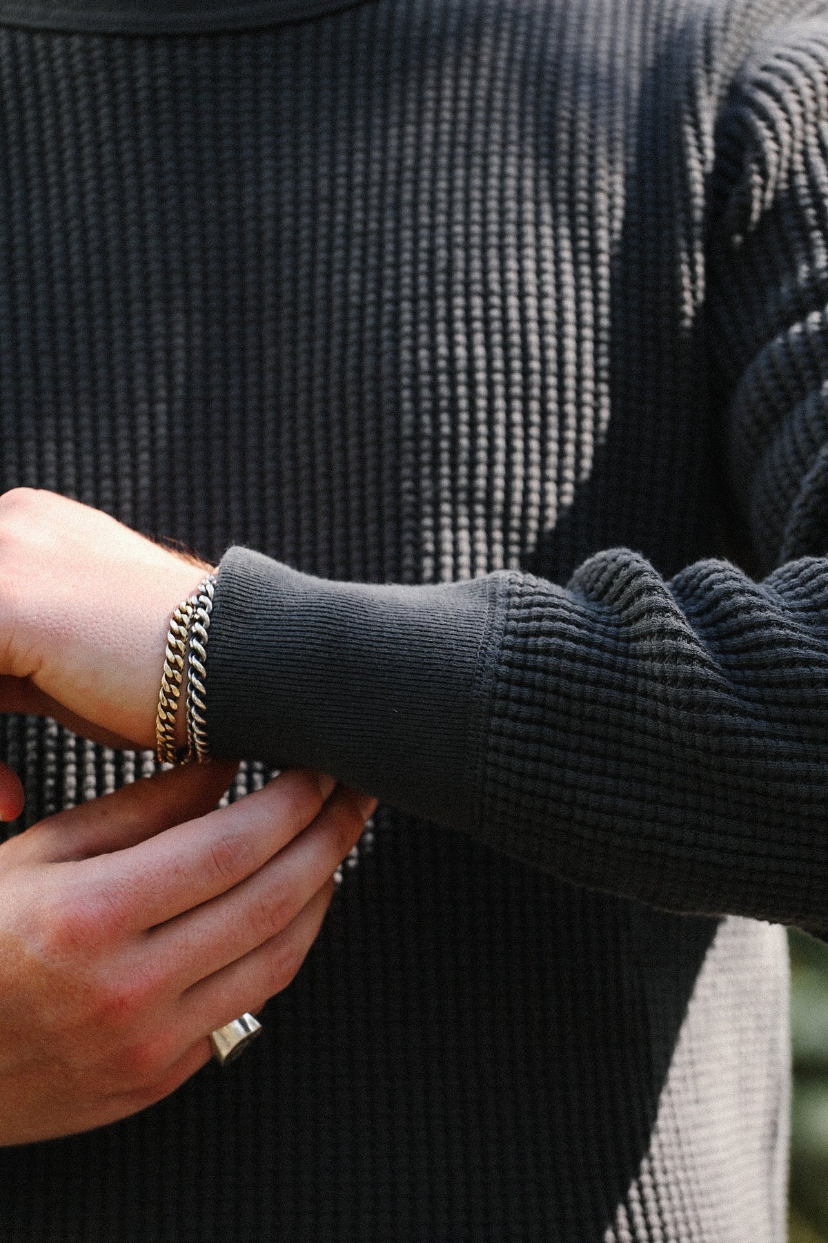 A close-up of a person wearing a dark charcoal gray ribbed knit sweater, showing the cuff of the sleeve and a gold chain bracelet on the wrist.