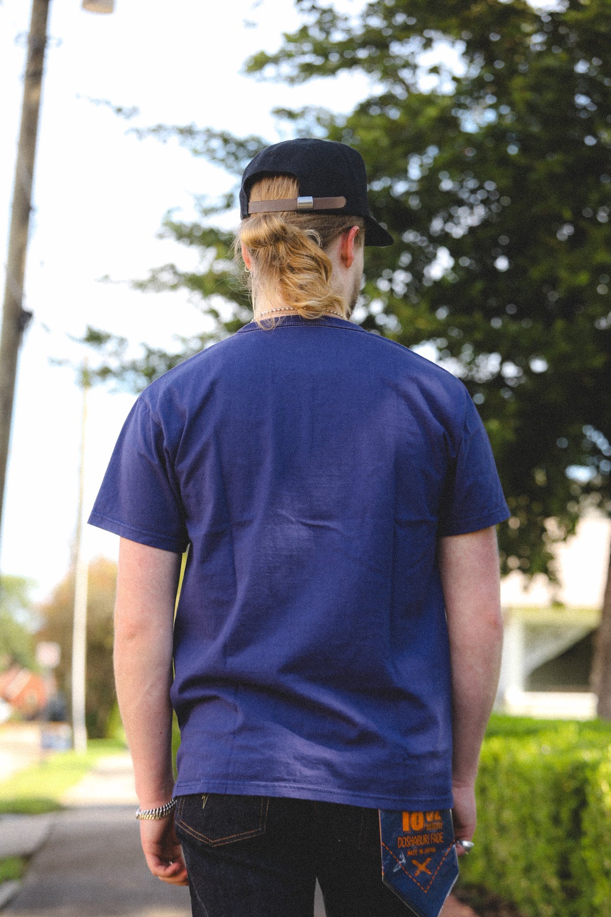 A person viewed from behind wearing a dark blue crewneck t-shirt, a black baseball cap, and dark jeans, standing outdoors on a path with trees in the background.