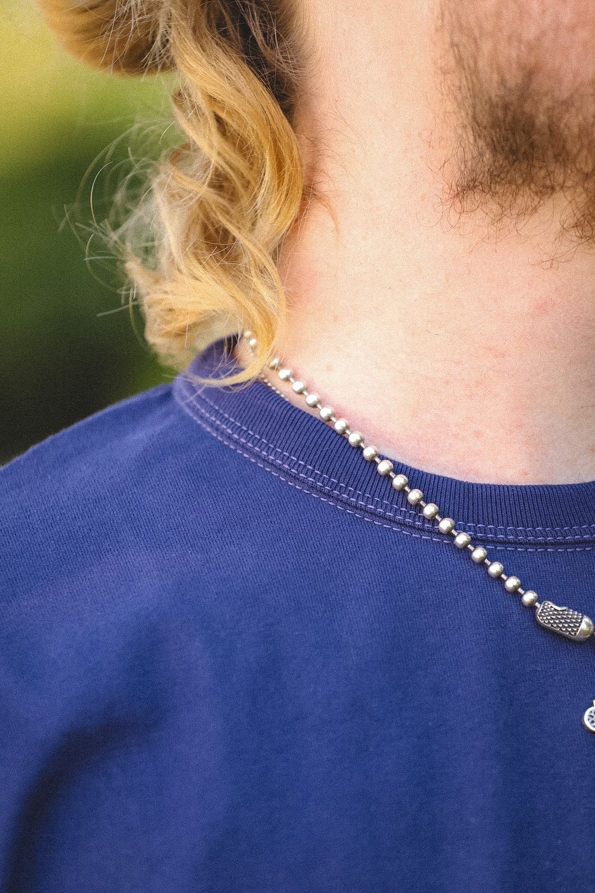 A close-up lifestyle shot of a person wearing a silver ball chain necklace with a textured metal pendant, paired with a navy blue crewneck t-shirt.