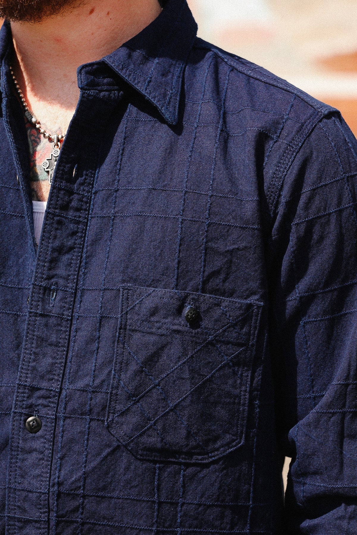 Close-up of a person wearing a dark navy textured button-up shirt with a chest pocket, showing part of the neck, necklace, and blurred warm-toned background.