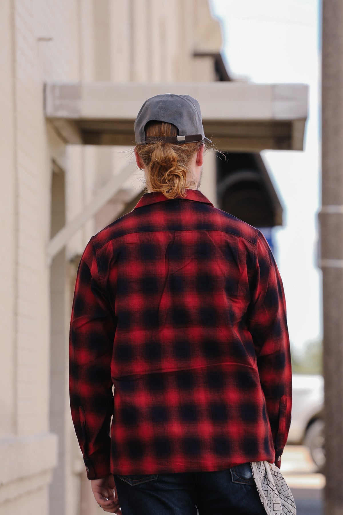 A person viewed from behind wearing a red and black buffalo plaid flannel shirt, dark jeans, and a gray baseball cap, standing outdoors under a building awning.