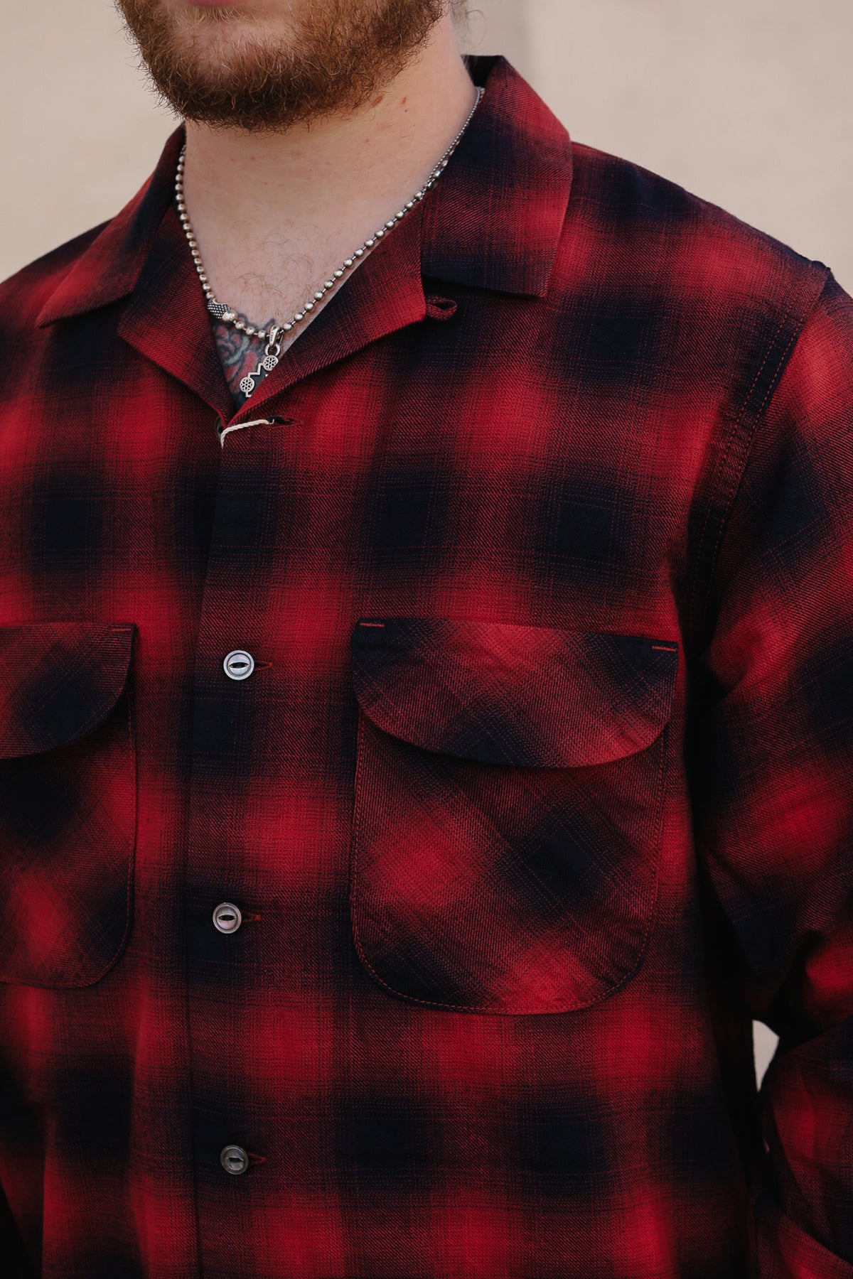 A close-up of a man wearing a red and black buffalo plaid flannel button-up shirt with two chest pockets and a silver chain necklace.