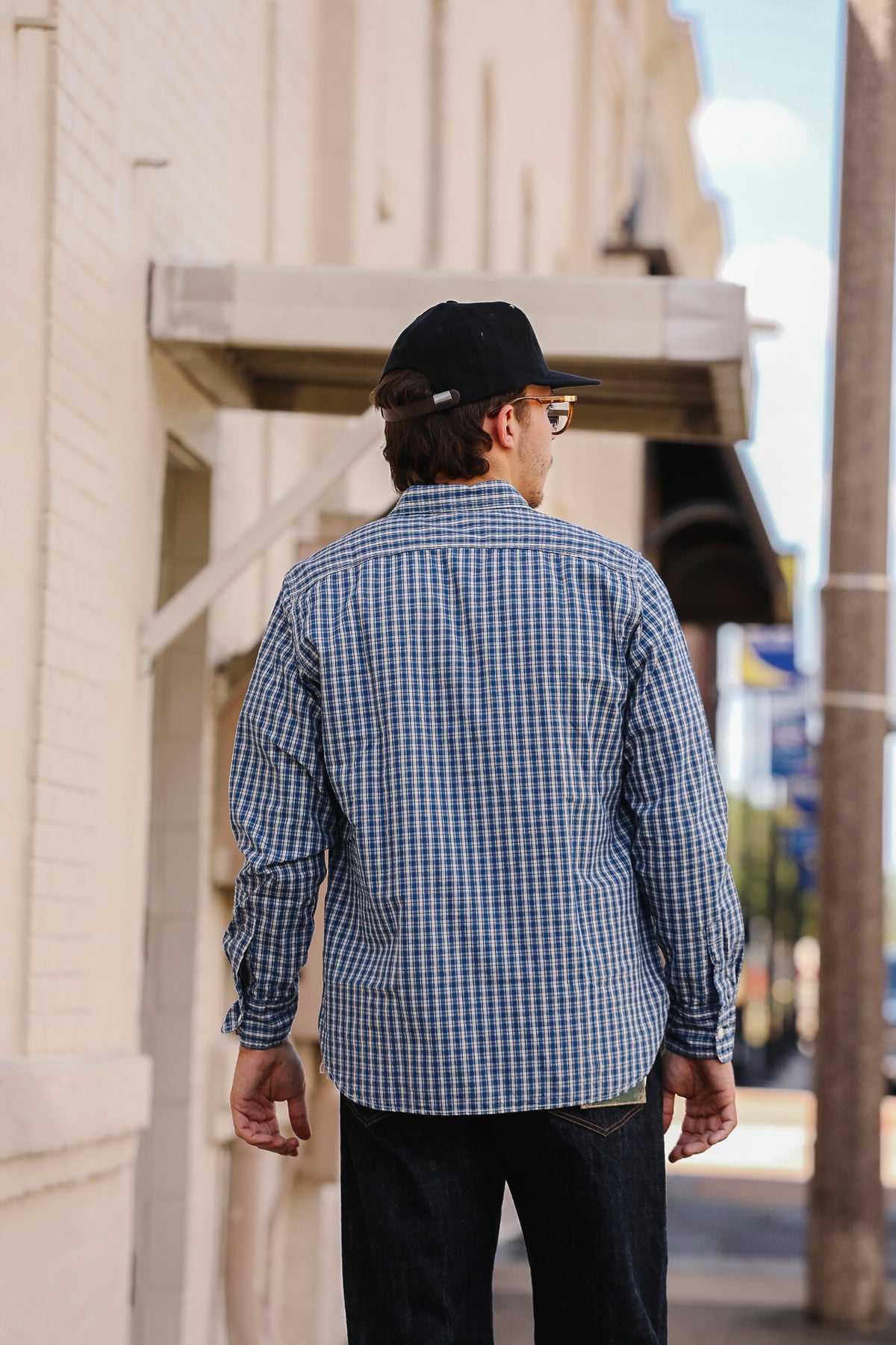 A man viewed from behind wearing a blue and white gingham long-sleeve button-down shirt, dark jeans, a black baseball cap, and sunglasses, standing on a city street next to a beige brick building.