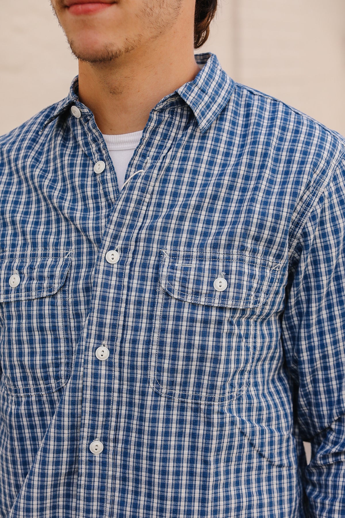 A close-up of a man wearing a blue and white plaid button-down shirt with two chest pockets, layered over a white undershirt.