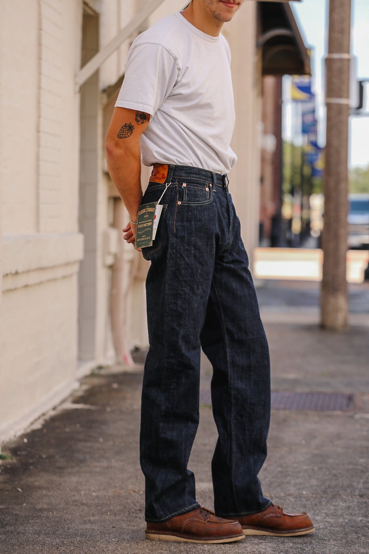 A man wearing dark indigo denim jeans with a brown leather belt and brown leather work boots, standing on a city sidewalk next to a white building.