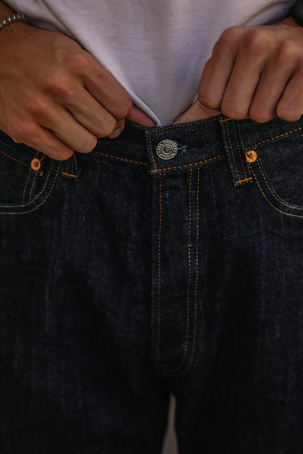 Close-up of a person wearing dark indigo denim jeans with orange contrast stitching, pulling the waistband down with both hands.