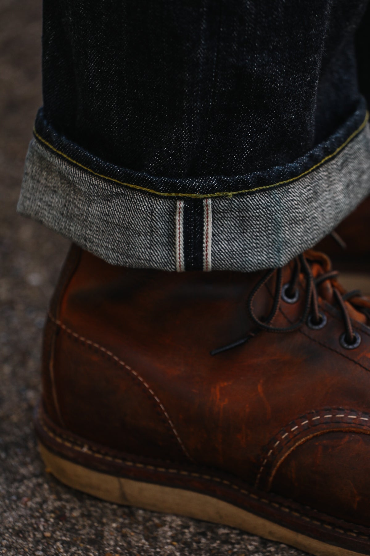 A close-up shot of a person's ankle wearing a dark indigo pair of jeans with a rolled cuff and a brown leather lace-up boot on a textured gray pavement background.