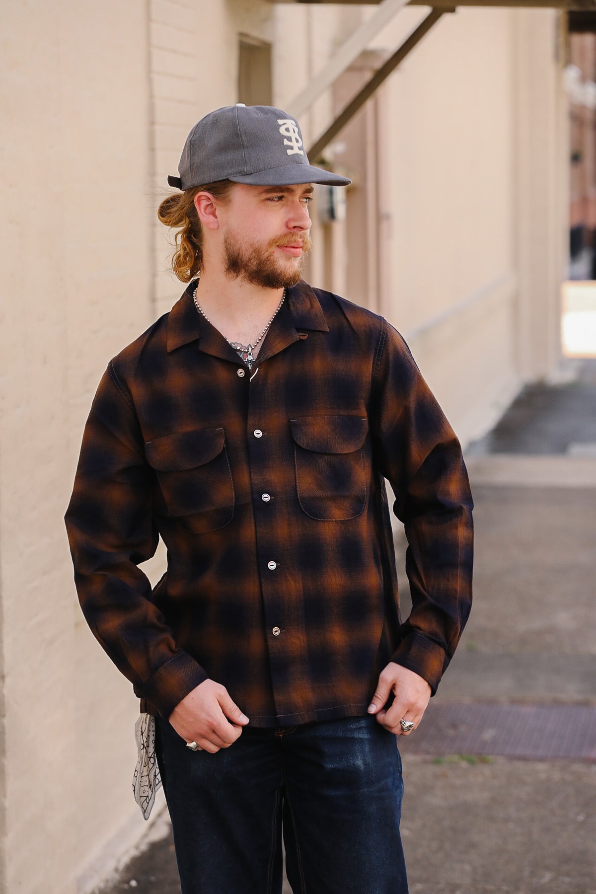 A man wearing a dark brown and black plaid flannel button-up shirt, dark blue jeans, and a gray baseball cap with a logo, standing outdoors against a beige building wall.