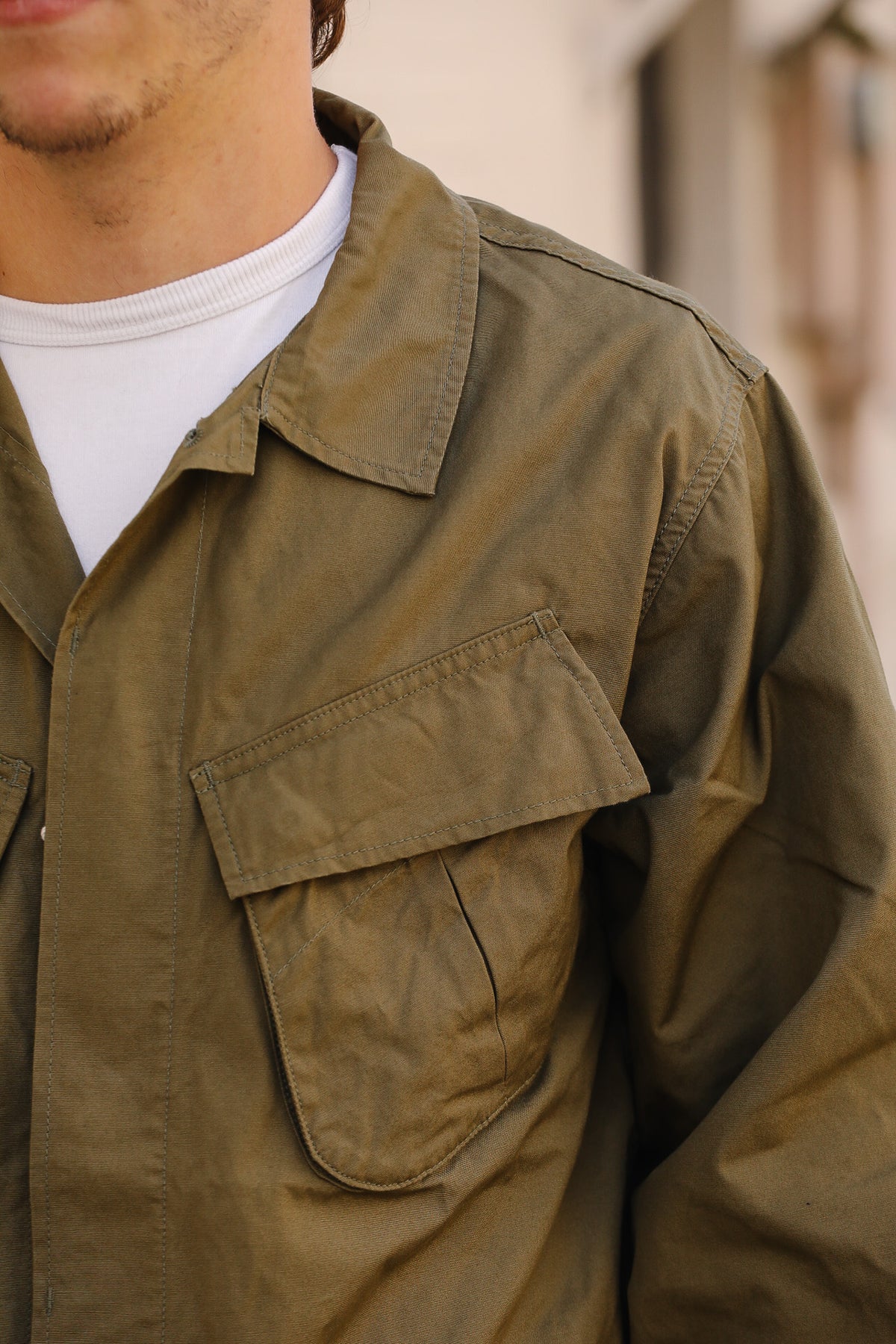 Close-up of a person wearing an olive green utility field jacket over a white t-shirt, with a softly blurred outdoor background.