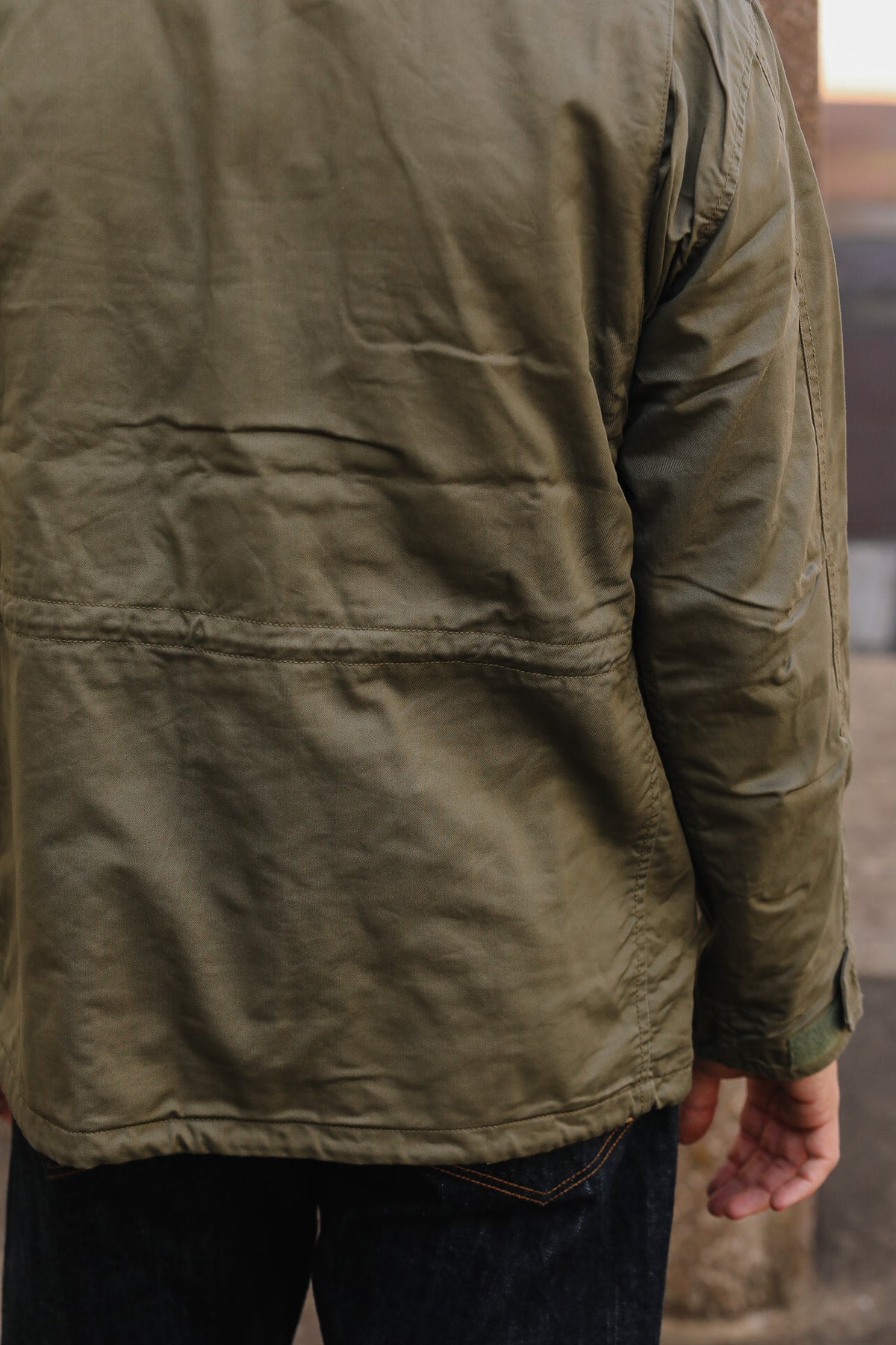 Close-up back view of a person wearing an olive green utility jacket with dark blue jeans, set against a blurred outdoor background.