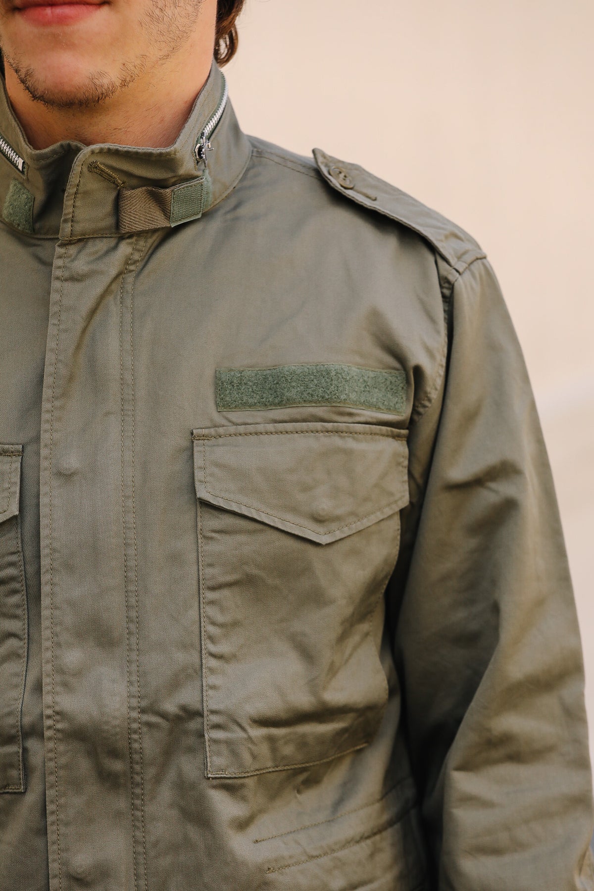 Close-up of a person wearing an olive drab military-style jacket, showing the chest pocket with a green velcro patch and collar detail.