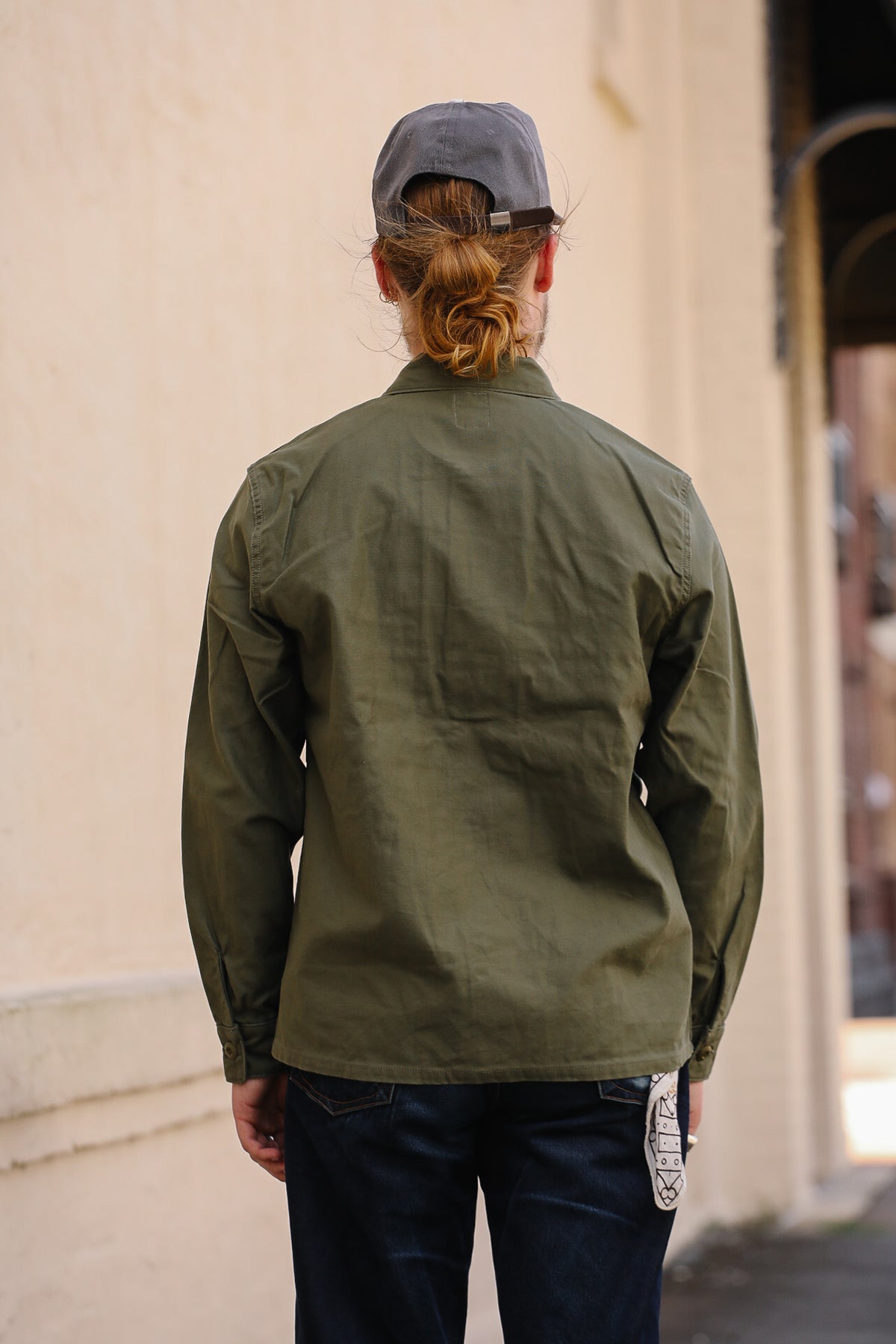 A person viewed from behind wearing an olive green utility jacket, dark blue jeans, and a gray baseball cap, standing against a beige wall.