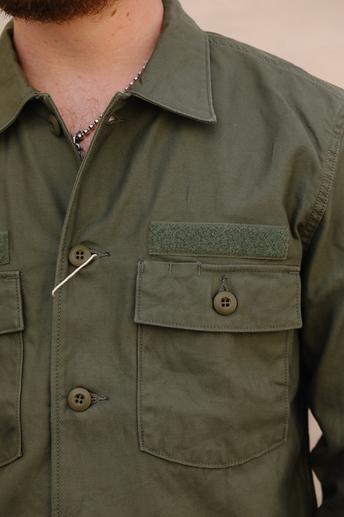 Close-up of a person wearing an olive green military-style button-up shirt with chest pockets, a buttoned collar, and a hook-and-loop flap detail above one pocket.