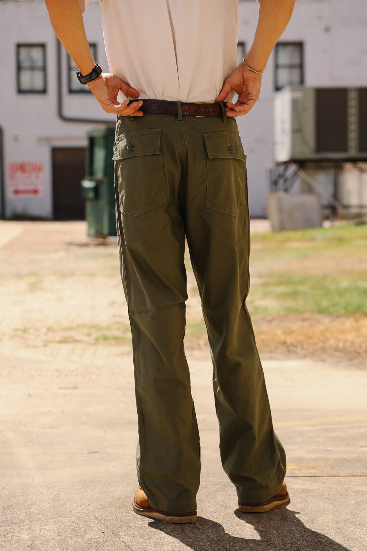 A person shown from the back wearing olive green cargo pants, a white shirt, and brown shoes, standing outdoors on a paved surface with a building in the background.